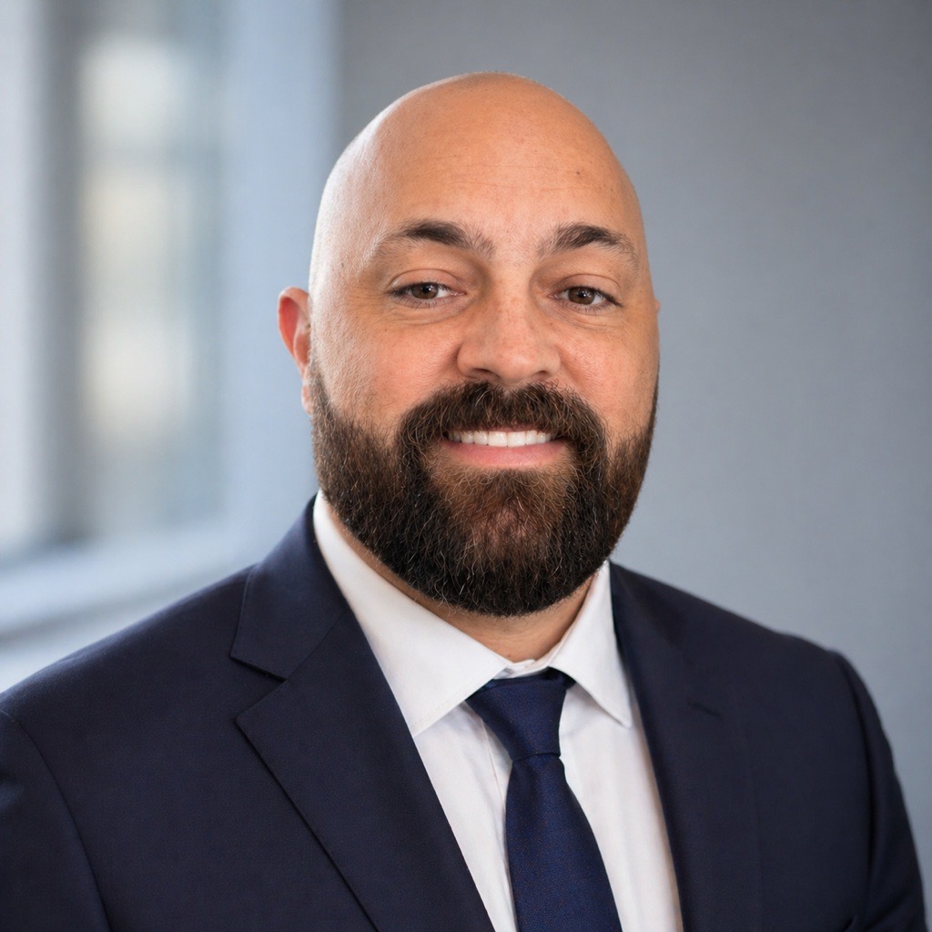 Professional headshot: bald man with a full beard in a navy suit and blue tie, smiling at the camera.